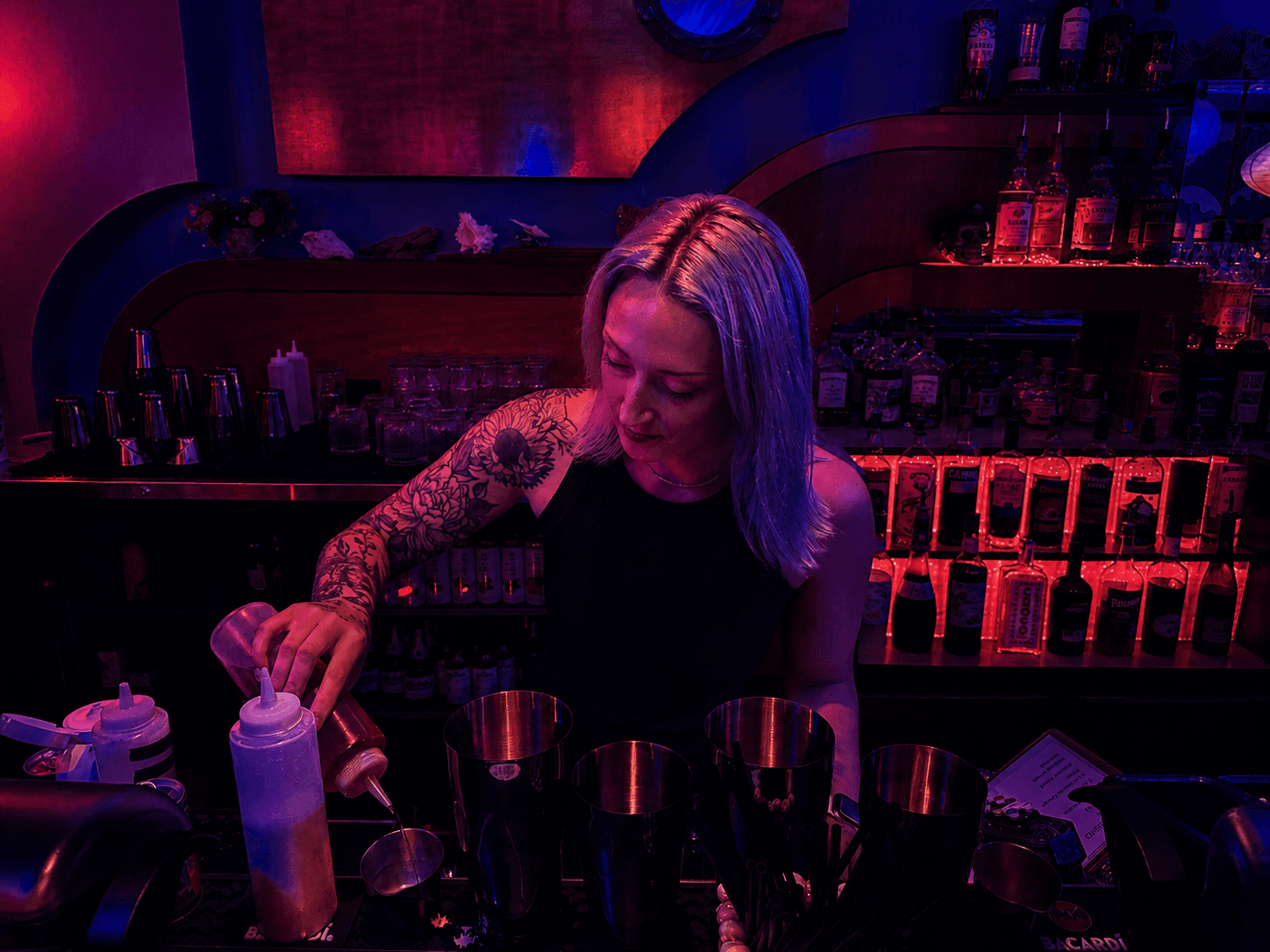 Tattooed bartender pouring ingredients into a shaker in a moody, neon-lit bar setting.