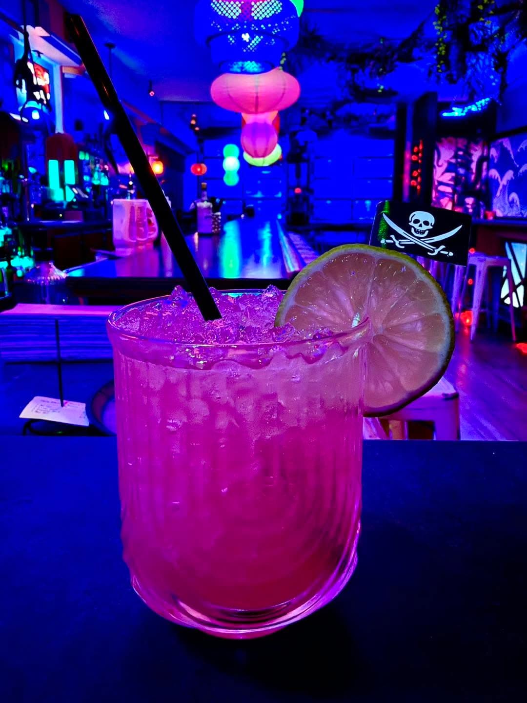 Vibrant pink cocktail with lime and pirate flag garnish in a neon-lit tropical bar.