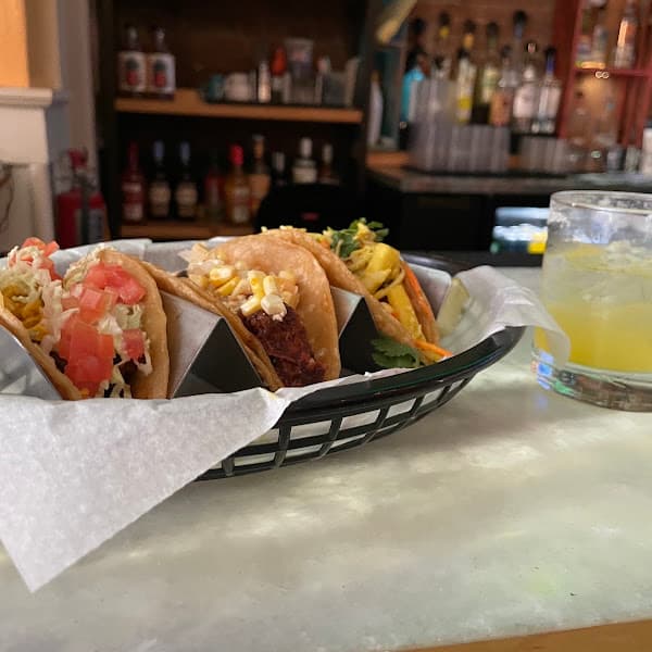 Three gourmet tacos with various toppings in a basket next to a yellow cocktail.