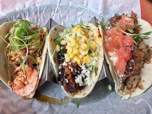 Three gourmet tacos with meat, corn, grapefruit, and microgreens served in a metal taco holder.