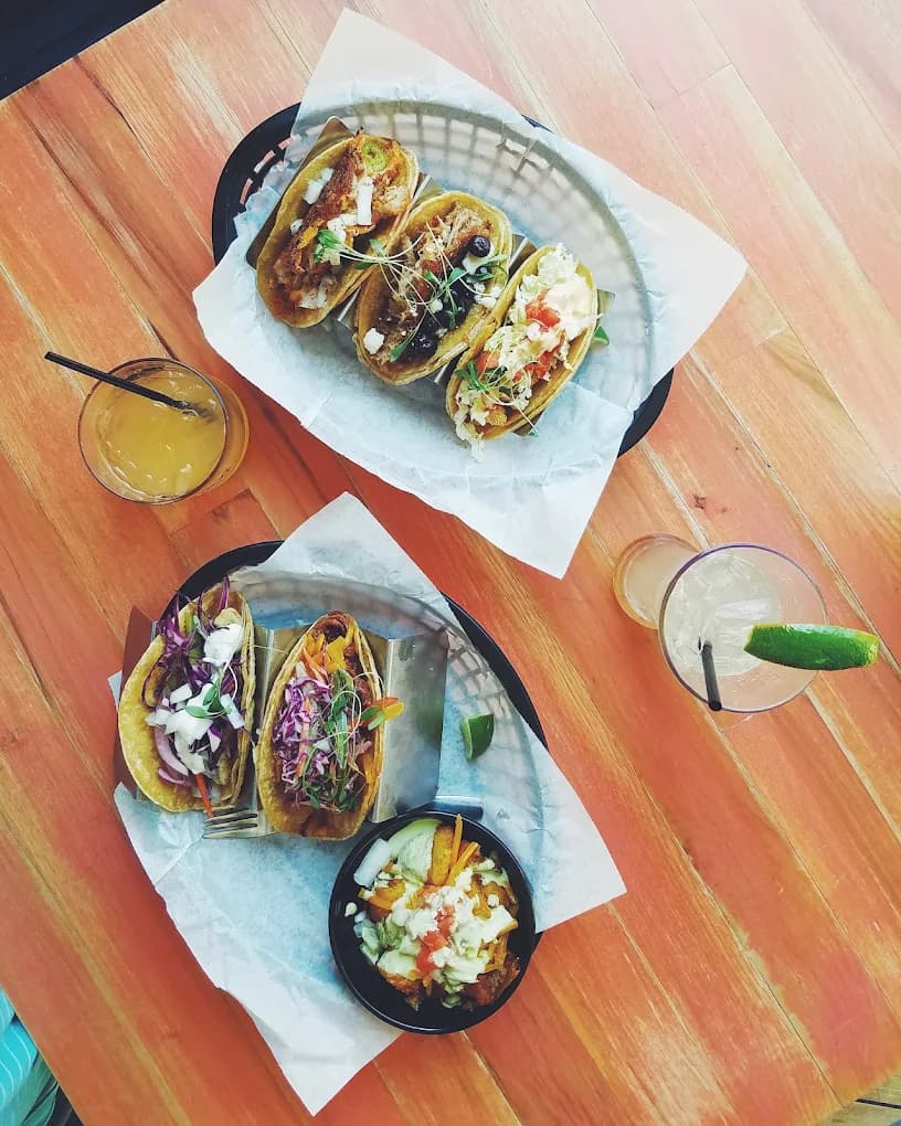 Overhead view of assorted colorful tacos and two drinks on a wooden table.